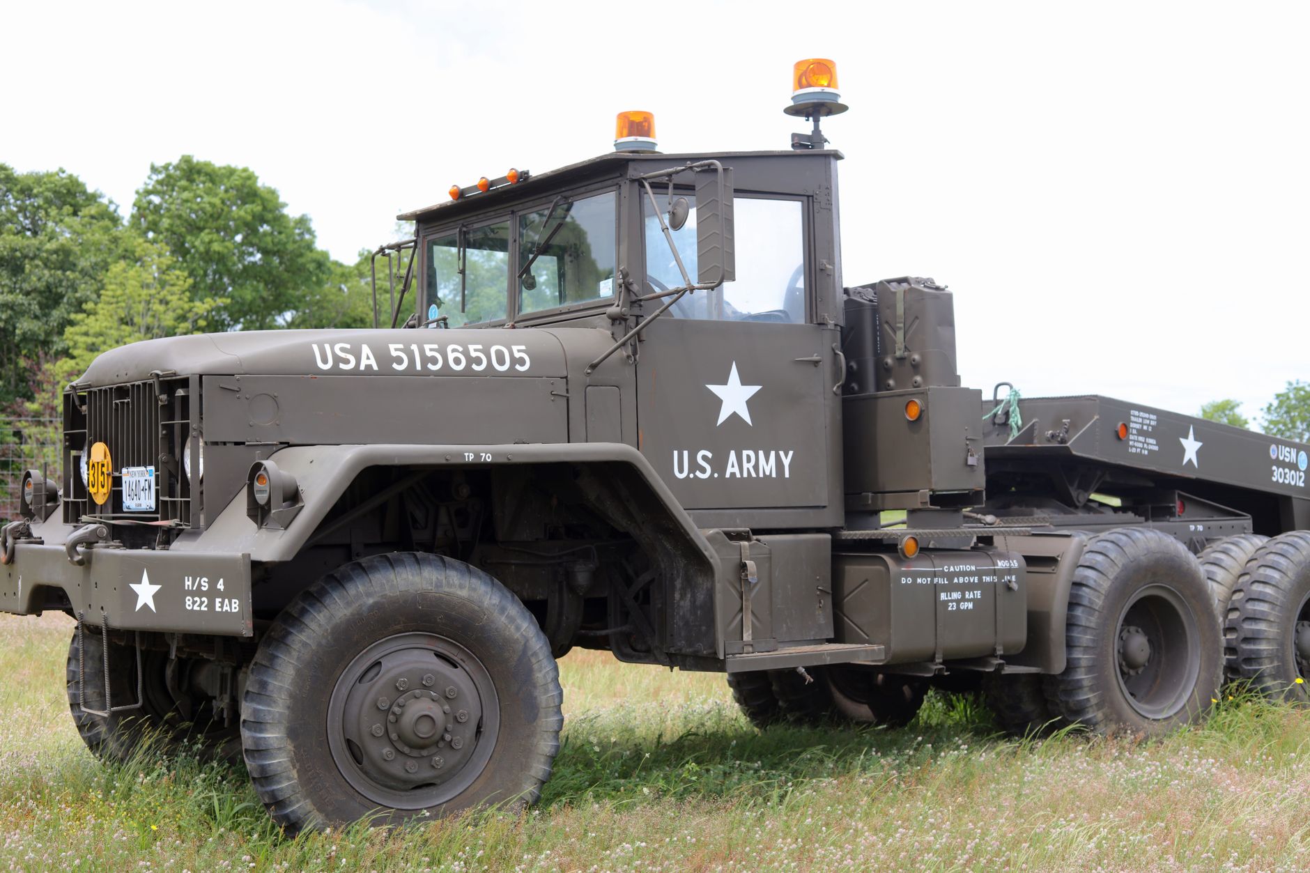 A weathered olive-drab military transport truck parked in a rural field, fitted with a small stovepipe chimney and curtained windows